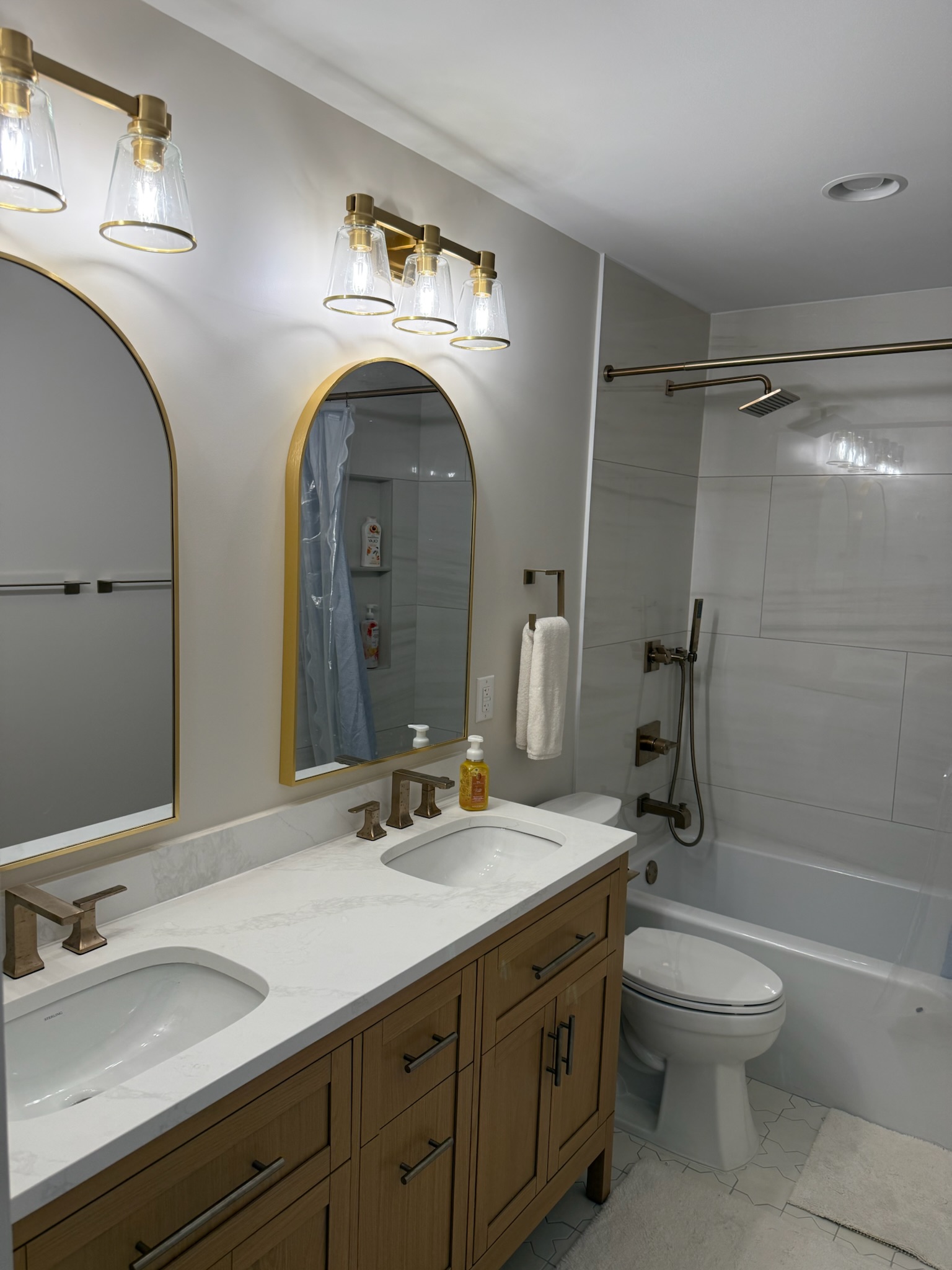 Warm transitional bathroom with oak double vanity, twin gold arched mirrors, marble-look quartz, and brushed gold fixtures in Upstate SC