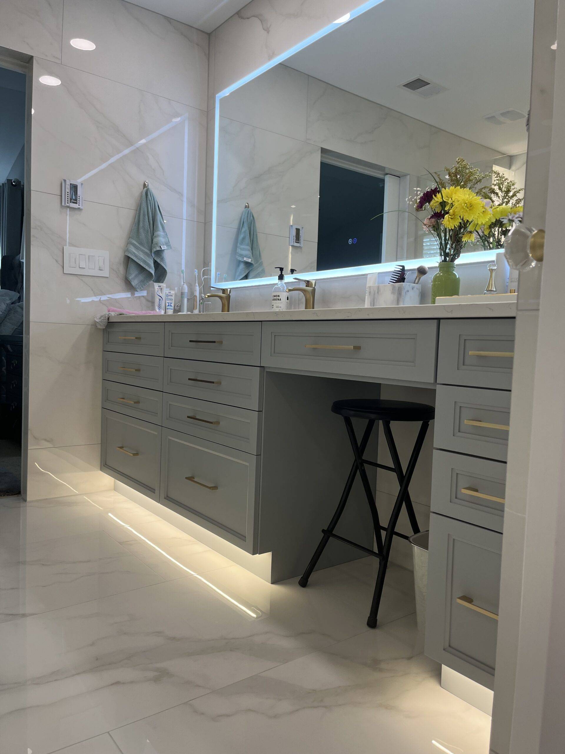 Modern bathroom interior featuring a sleek gray vanity with gold hardware, illuminated mirror, floral arrangement, and stylish lighting, showcasing Upstate Bath Renewal's remodeling expertise.
