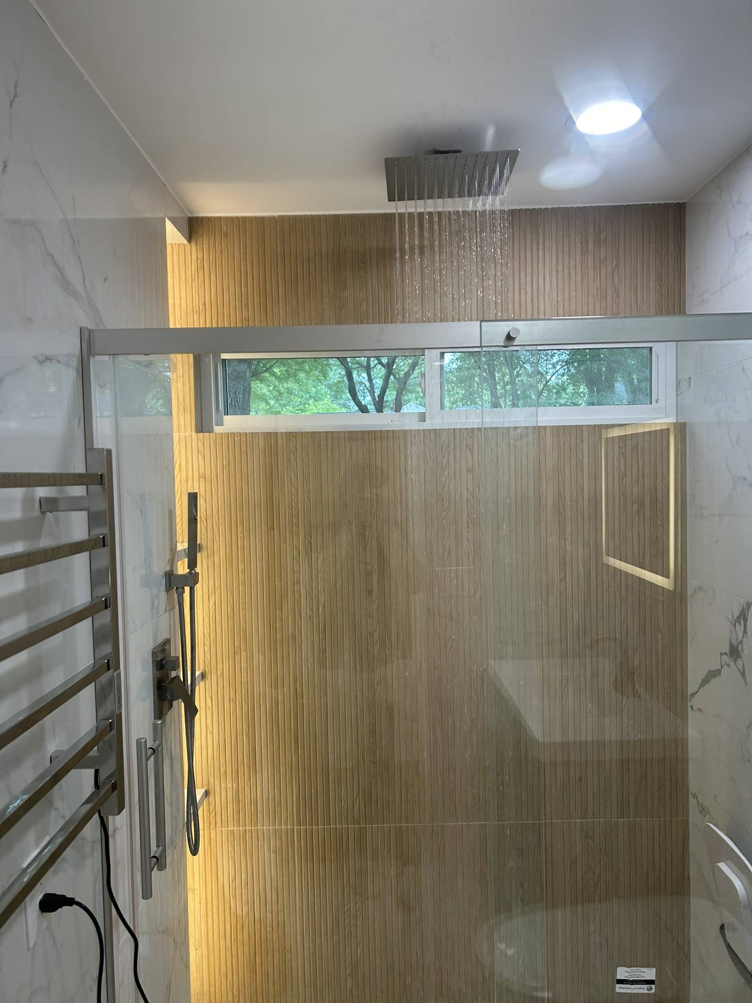 Modern shower featuring contemporary rainfall showerhead, sleek glass enclosure, warm wooden accent wall, and bright lighting, reflecting high-quality bathroom renovation by Upstate Bath Renewal.